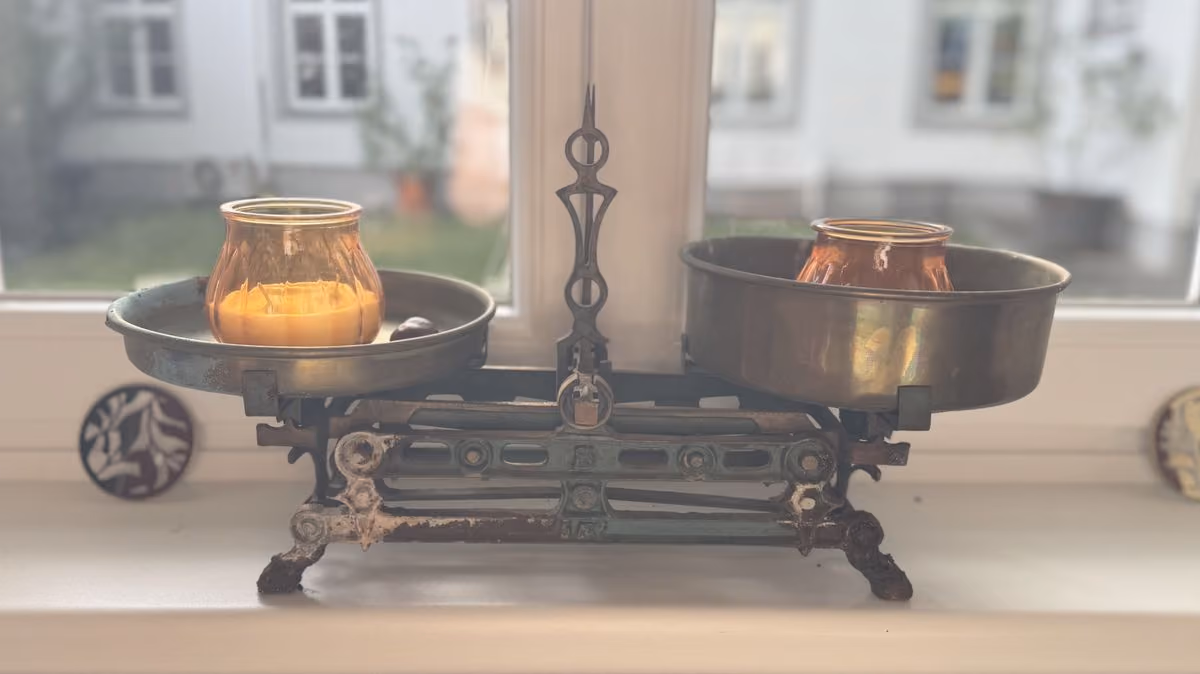 Eine antike Waage auf einer Fensterbank, auf jeder Schale ein bernsteinfarbenes Glas, als Sinnbild für innere Balance.